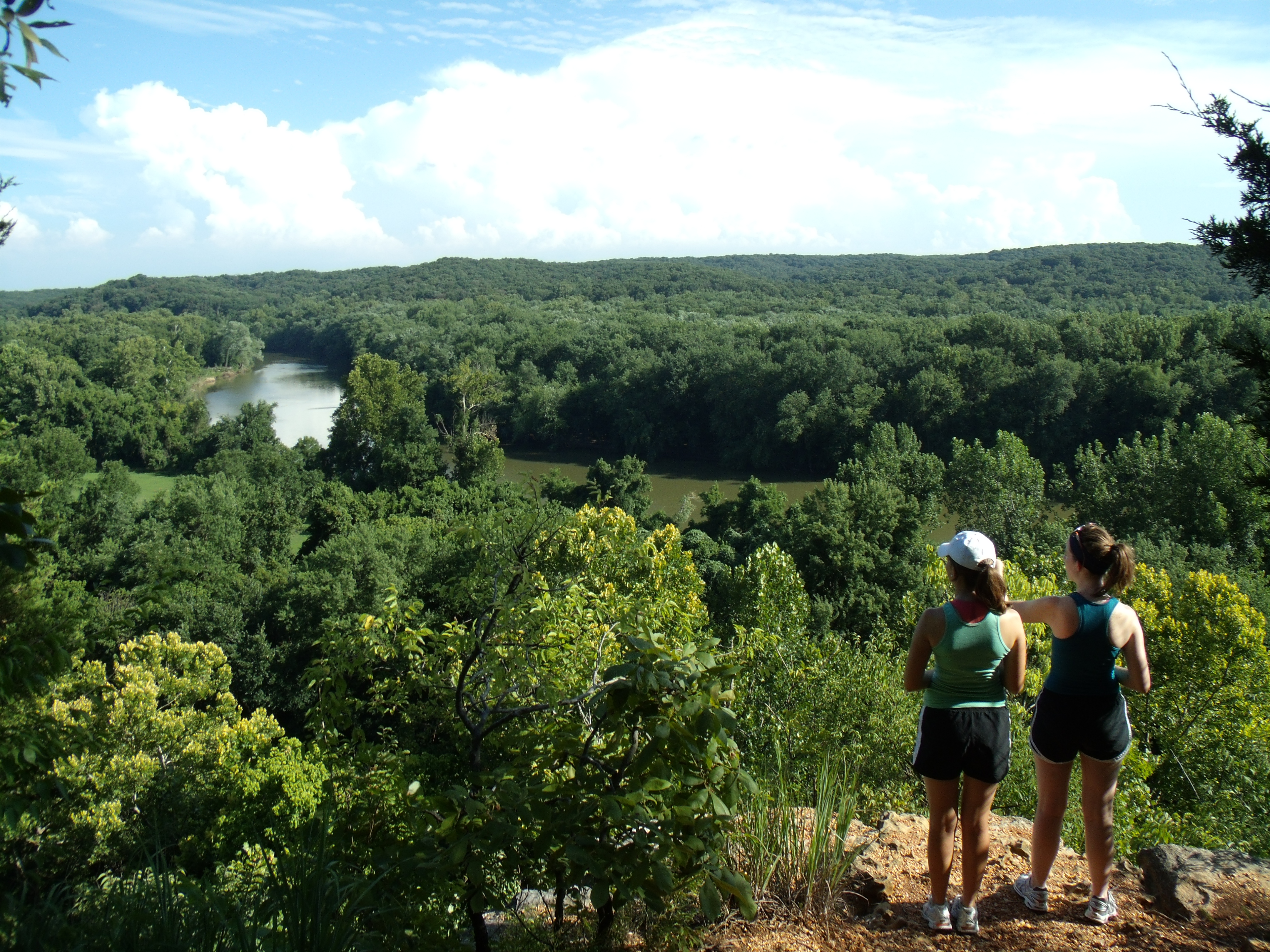 two women on a cliff looking over a forest with a river cutting through it