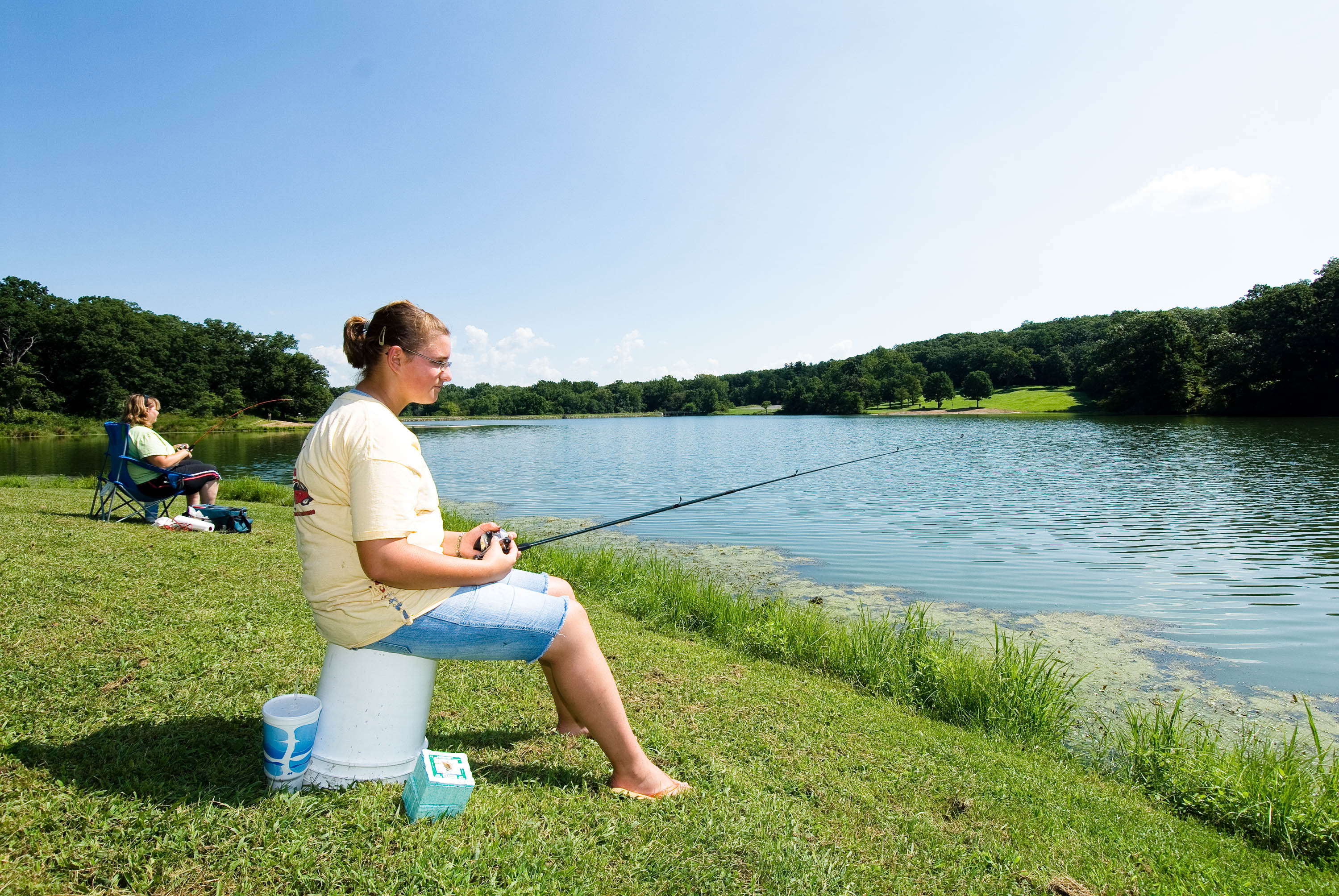 a woman sitting on a plastic bucket fishing into a body of water