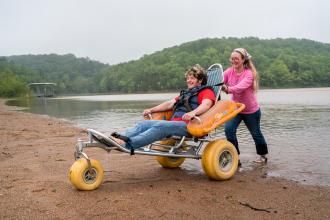 Photo showing person in beach chair being pushed by another person at a beach.