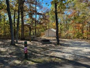 A wooden campsite post with an R and the number 45 stands in front of a fire ring and a wooden picnic table in a shady area
