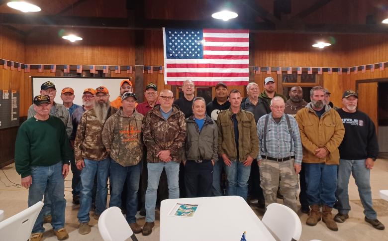 Twenty men stand in front of an American flag in a large room