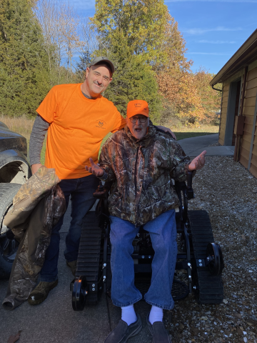 A man sits in a large motorized chair while another man stands beside him