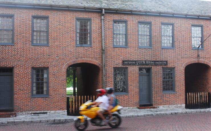 Two people on a yellow motorcycle driving past a brick general store