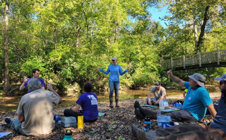 A woman speaks to a group of volunteers sitting on a creek bank