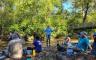 A woman speaks to a group of volunteers sitting on a creek bank
