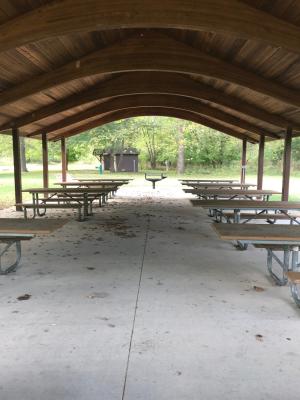 Two rows of picnic tables under the roof of the open shelter, with a grill, water fountain and restroom in the background