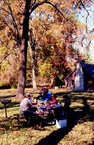 a family enjoys a picnic lunch at a picnic table under large shade trees