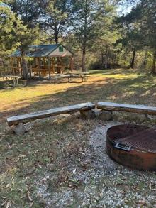 Metal fire pit and rugged wooden benches with wooden picnic tables and open shelter in the background