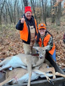 A veteran and a guide, both in camouflage and hunter orange pose with a harvested buck in the woods.