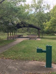 A water fountain stands on the sidewalk that leads to the open picnic shelter